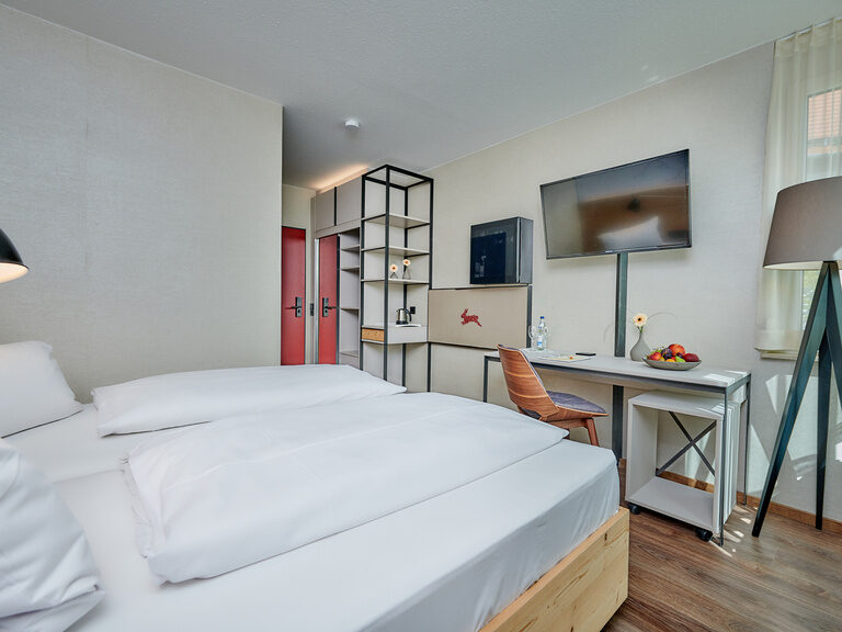 Modern eingerichtetes Hotelzimmer mit Doppelbett, Schreibtisch, Wandregal, Flachbildfernseher und großer Stehlampe neben einem Fenster.